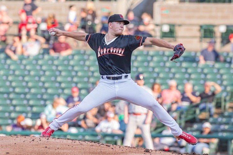 Pitcher shown throwing a ball. He is wearing a black ASL jersey, baseball cap, grey pants, and red shoes.