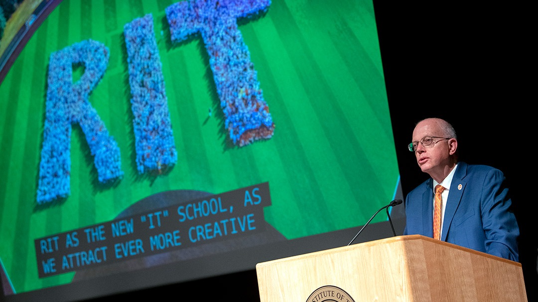 RIT President Munson stands at podium and addresses RIR community. A slide with students spelling out RIT is behind him.