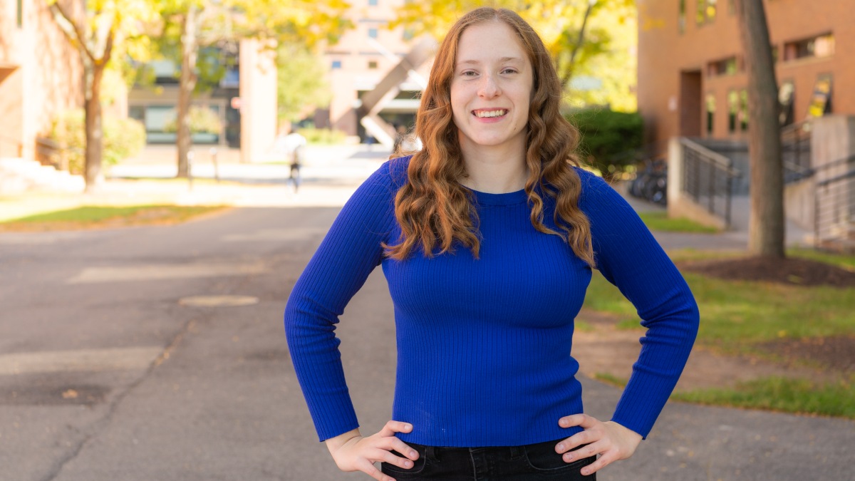 Woman with long red hair wearing a bright blue top stands with arms on hips.