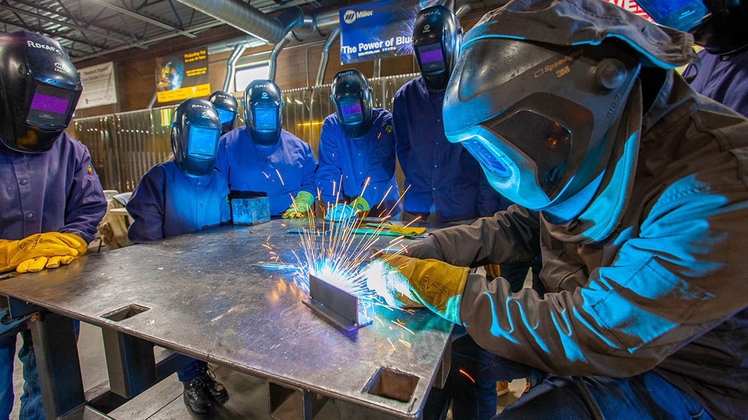 Eight people wearing welding gear stand around a table. One person is making sparks. 