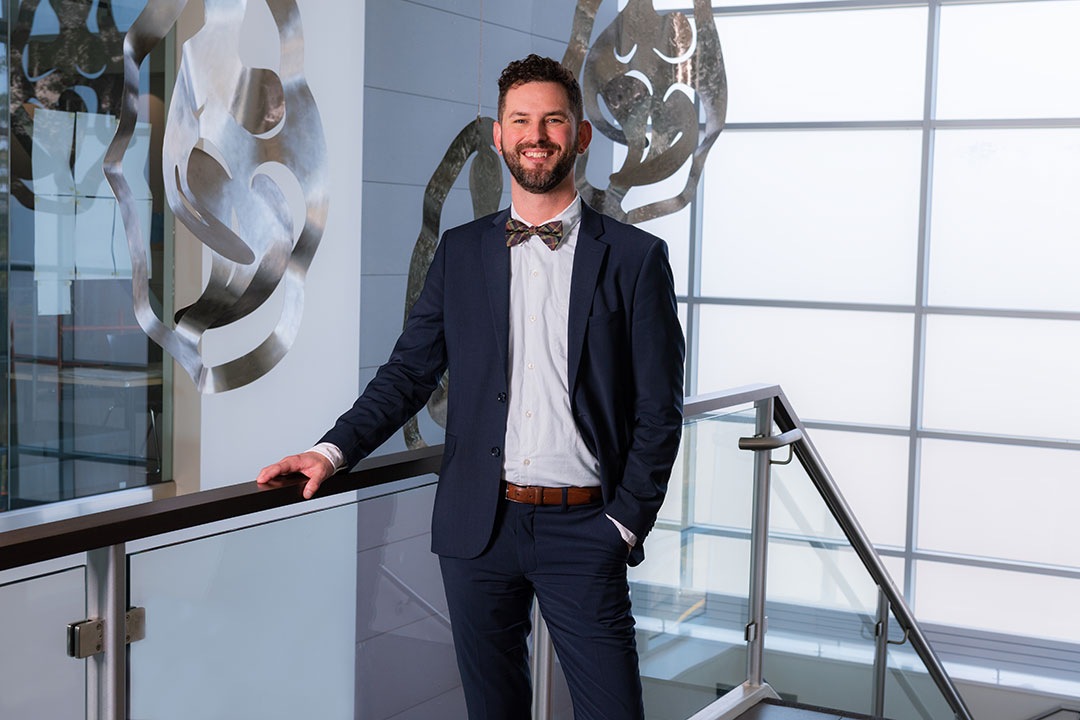 Male standing with short dark hair and a beard and mustache is wearing a blue suit, white shirt and multi-colored bow-tie.