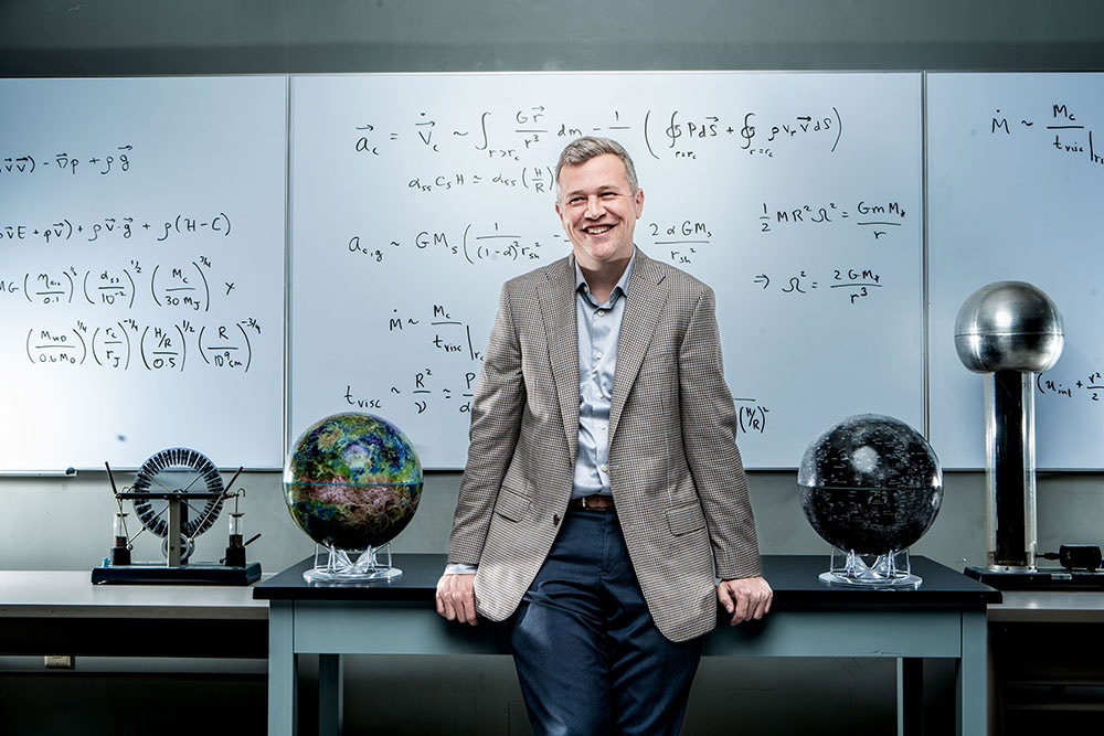 Male professor wearing a tweed jacket, light blue shirt and blue pants is smiling and leaning against a lab table. In the background is a white board with mathematical equations.