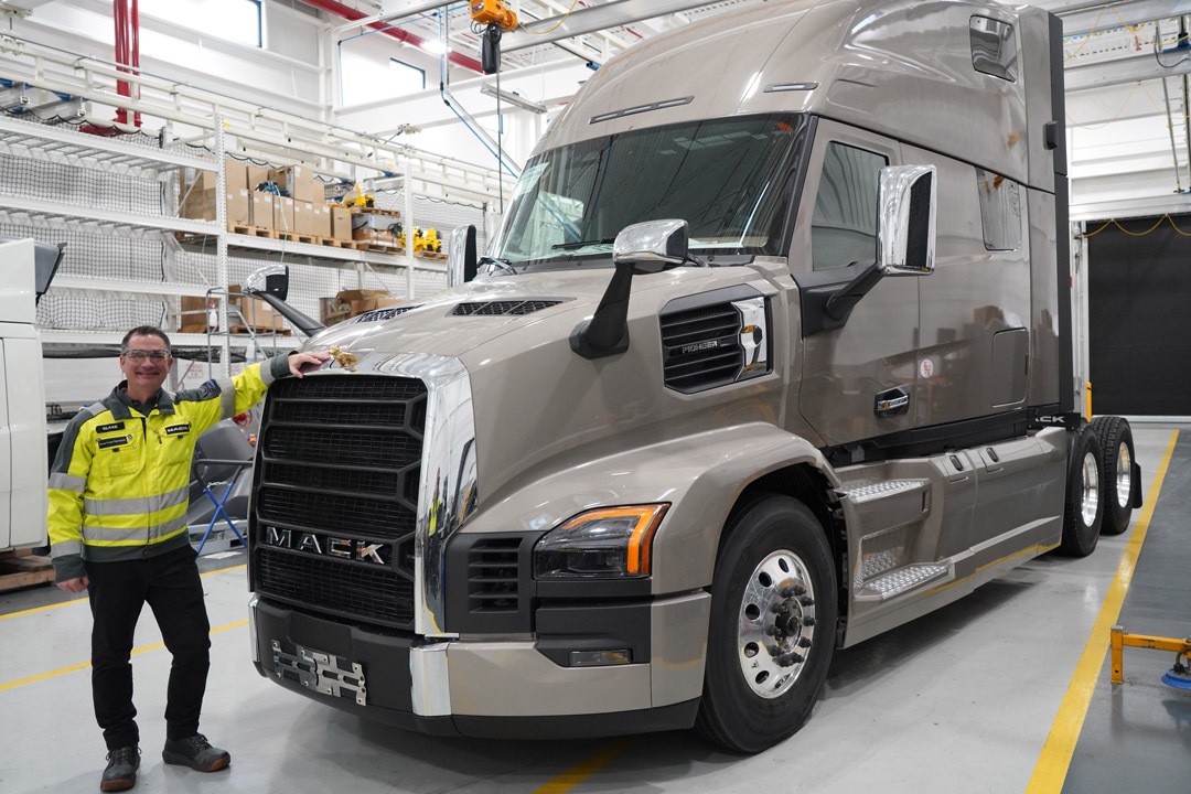 a man in a yellow safety jacket stands in front of a mack truck