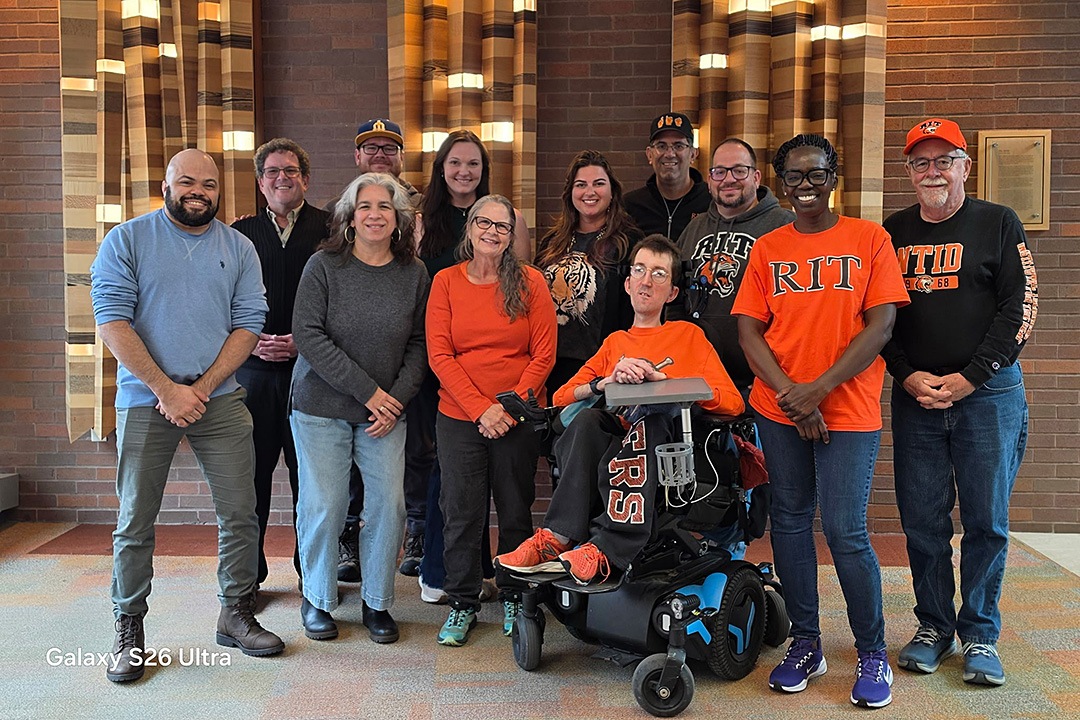 A group of eleven diverse individuals posing together indoors against a brick wall with decorative light panels. Several group members are wearing Rochester Institute of Technology (RIT) and NTID-branded orange and black apparel, including shirts, hoodies, and hats. In the center, a man in an orange shirt sits in a motorized wheelchair. Everyone is smiling for the camera.