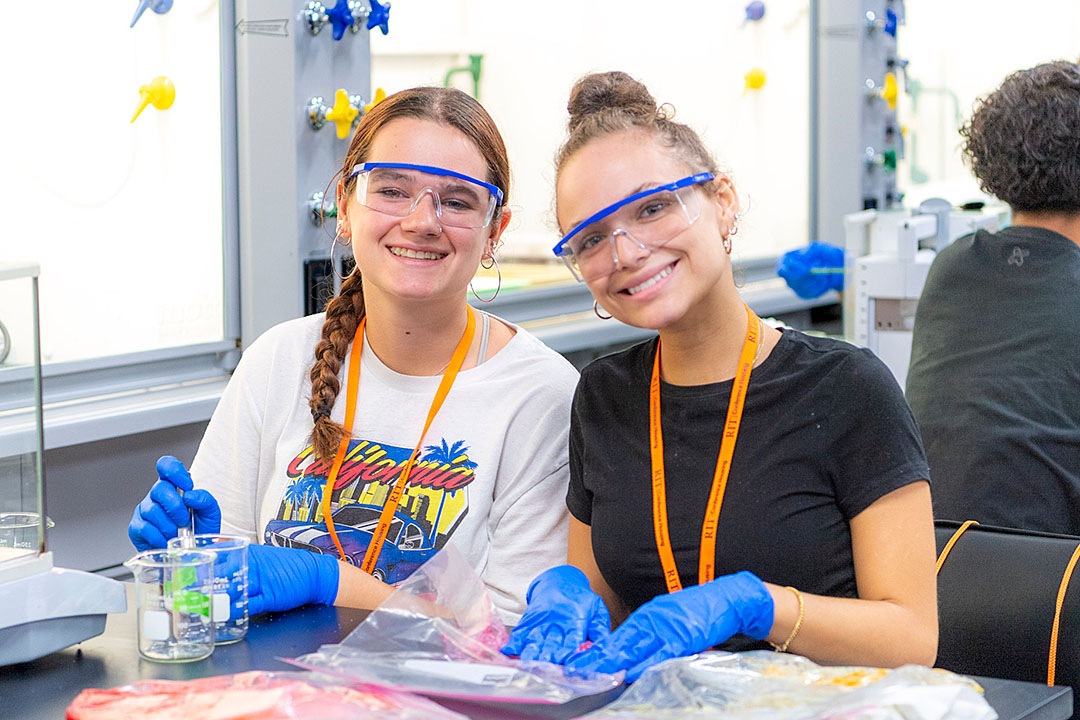 Two individuals wearing safety goggles and gloves working on a science experiment in a laboratory setting.