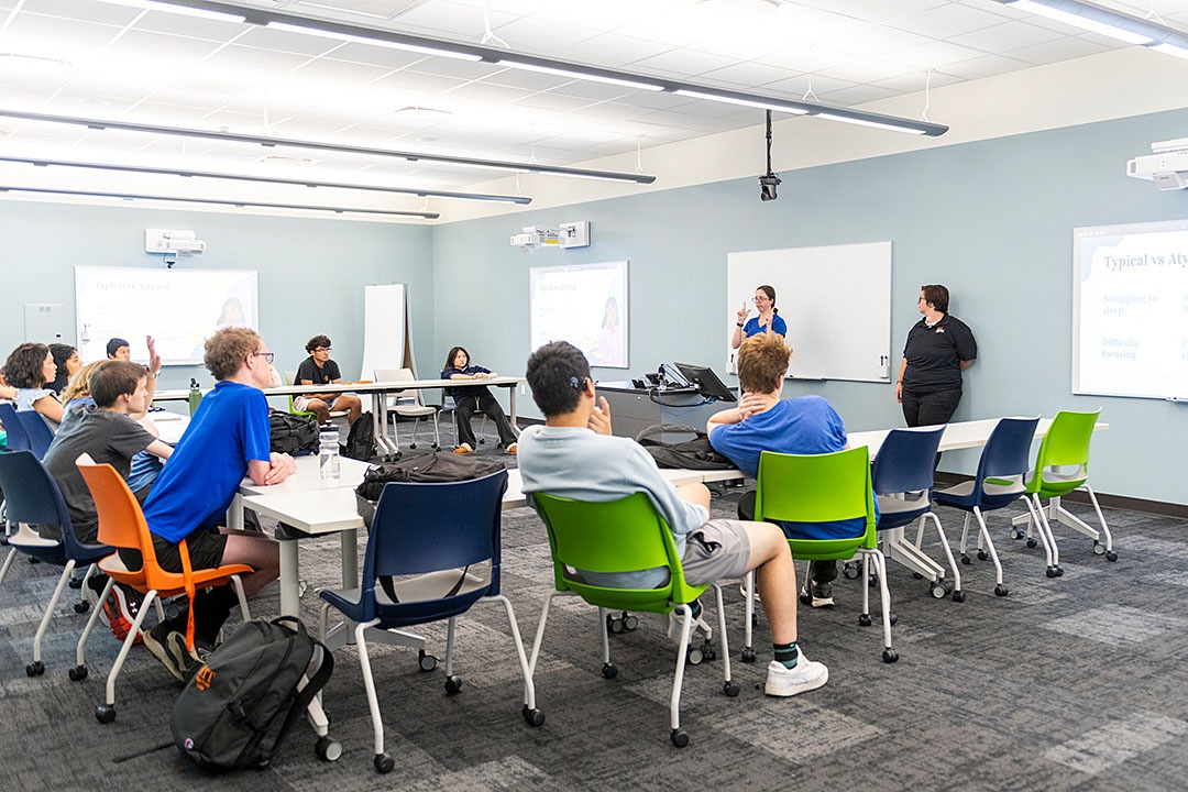 A bright, modern classroom where a diverse group of students sits at white tables. An instructor stands at the front by a whiteboard using American Sign Language (ASL) while another staff member stands nearby.