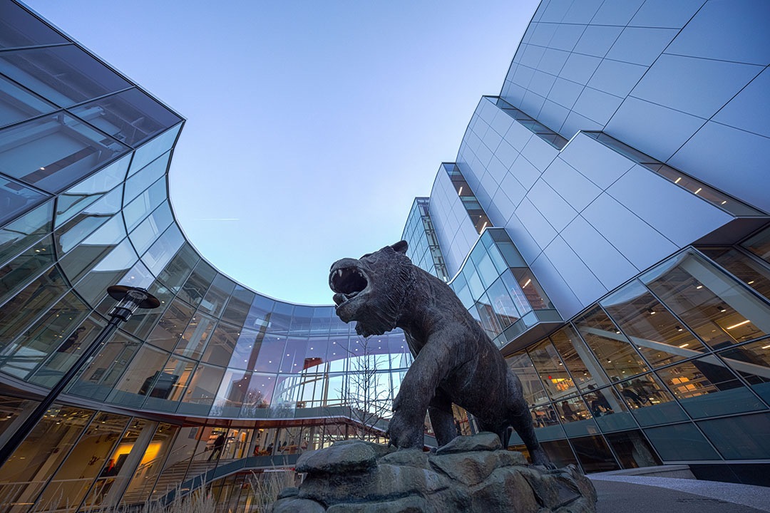 A large bronze tiger statue roaring in a modern campus courtyard surrounded by curved glass and steel buildings under a clear blue sky.