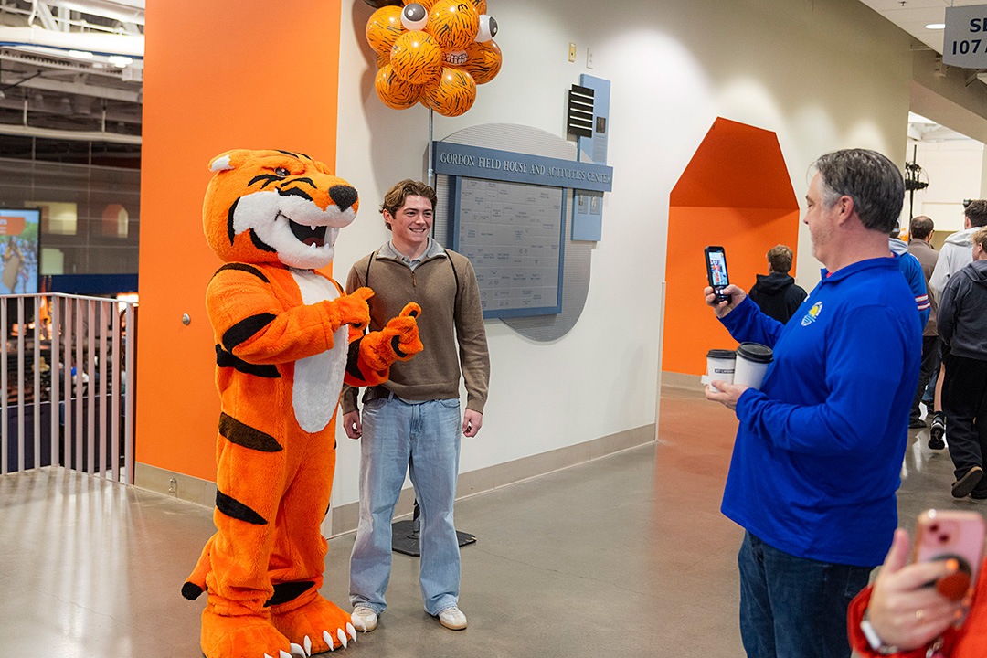 Inside the Gordon Field House and Activities Center, a perspective student poses for a photo with the Richie the Tiger mascot while an onlooker takes a picture with a smartphone. The scene features bright orange walls, tiger-striped balloons, and visitors walking through the open house event.