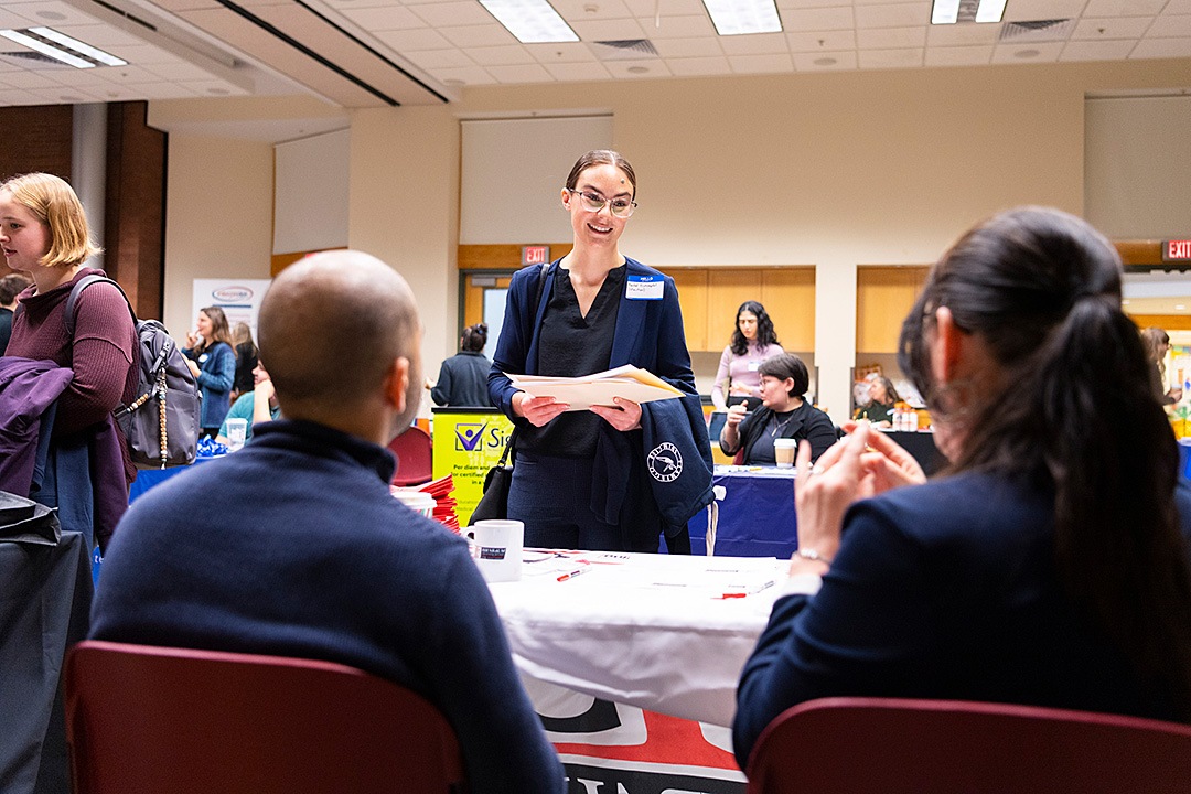 A smiling young woman holds a folder while speaking with two seated representatives at a busy career fair.