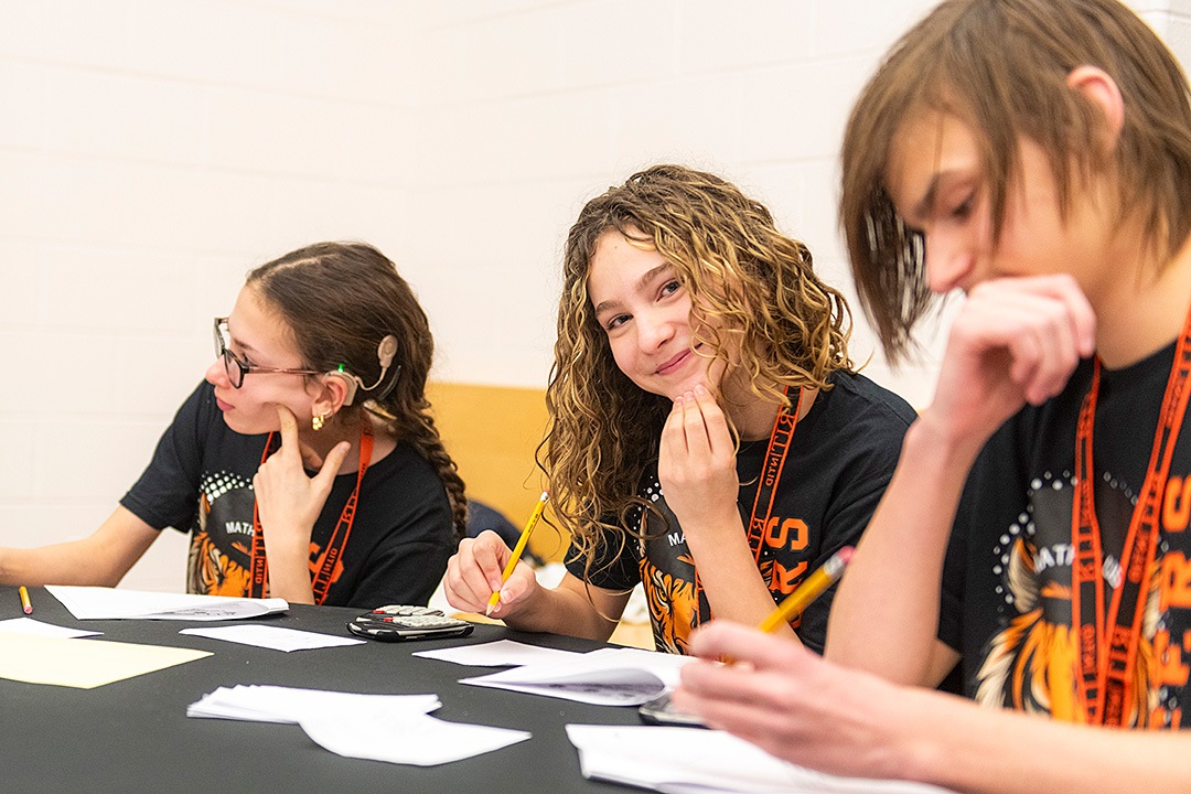 Three students wearing matching black RIT NTID tiger t-shirts sit at a desk with papers, pencils, and a calculator. The girl in the center smiles warmly at the camera, resting her chin on her hand. The girl on the left, who is wearing glasses and a behind-the-ear hearing device, looks off to the side, while the boy on the right looks down toward his desk.