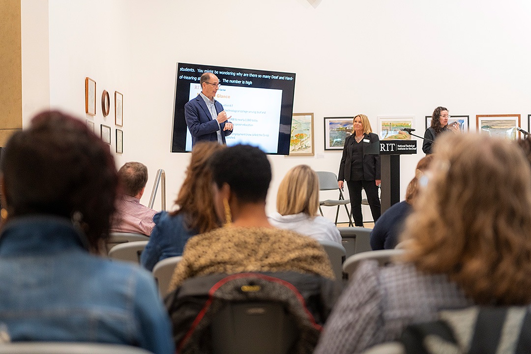 A man speaks and signs in front of a digital presentation screen to a seated audience inside a gallery space. Beside him, a woman stands behind a podium bearing the RIT National Technical Institute for the Deaf logo, while another woman to the far right provides sign language interpretation.