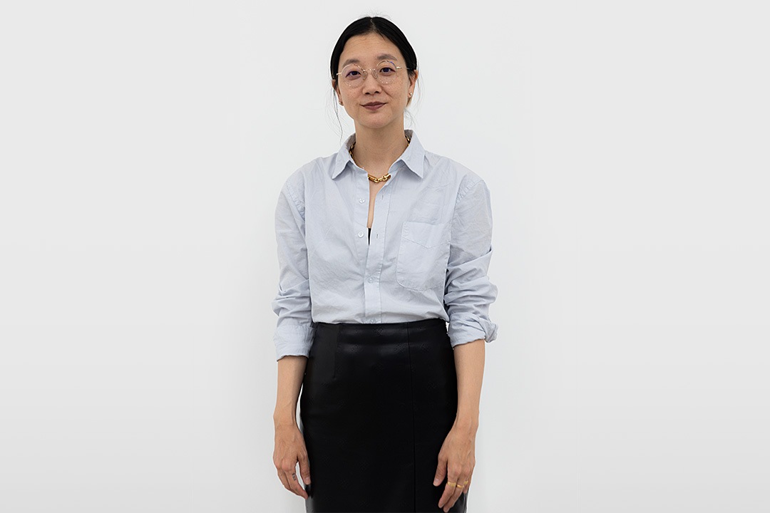 A portrait of Christine Sun Kim standing against a plain white background. She is looking directly at the camera with a subtle smile and wears round wire-rimmed glasses, a light blue button-up shirt, a black skirt, and a chunky gold chain necklace.