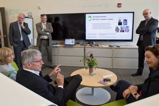 A group of people around 2 televisions talking while some sit and some stand