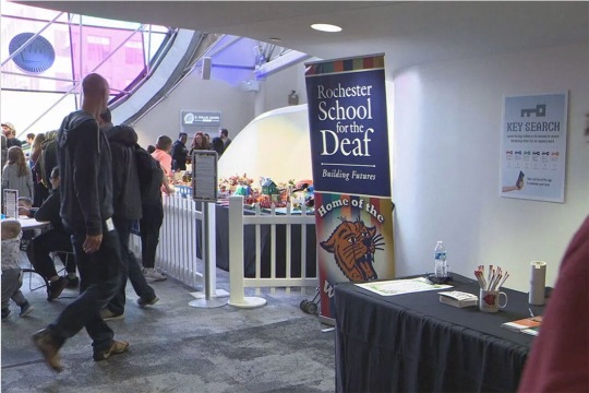 the interior of the Strong Museum is shown with people mingling on the left side of the frame and an informational table about the Rochetser School for the Deaf on the right.