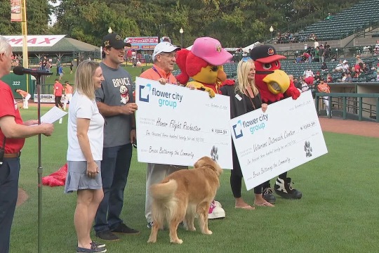 A group of people, including the Red Wings mascots and a golden retriever, stand on the baseball field holding two oversized checks from Flower City Group to local charitable causes.