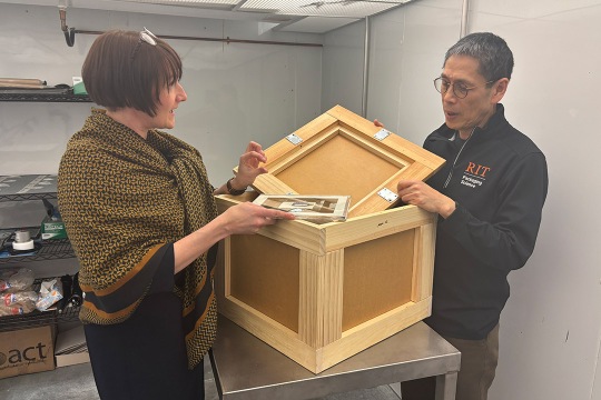 Two people examine a wooden box with hinged panels on a metal table. The woman holds a photo. The man wears glasses and a shirt with an RIT logo.
