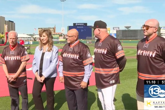 5 people stand on a baseball field wearing jerseys that spell Rochester in ASL sign.