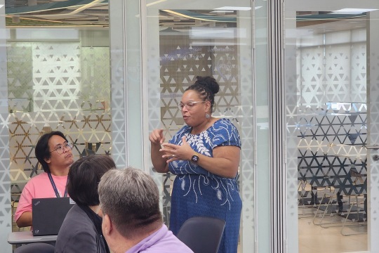 a woman in a blue and white dress speaks to other adults in a modern classroom environment.