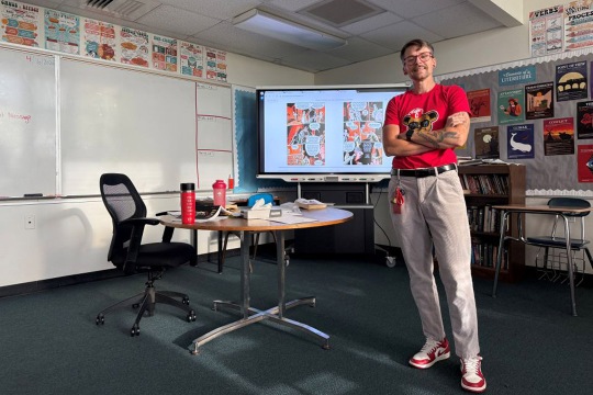 a tall person wearing khaki pants and a red shirt stands in a classroom with a whiteboard lit up with a lesson behind them.