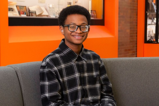 Person seated on a gray couch in a brightly lit space with an orange wall, wearing a black-and-white plaid shirt.