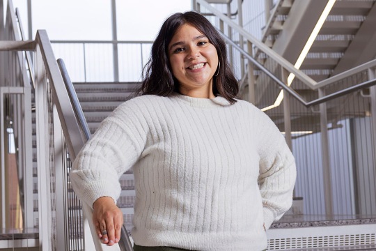 Person standing on an indoor staircase wearing a white knit sweater, with one hand resting on the railing.