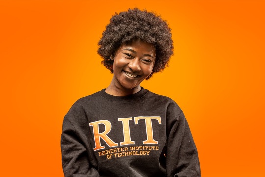 Person seated against an orange background wearing a black sweatshirt with “RIT Rochester Institute of Technology” printed on it.