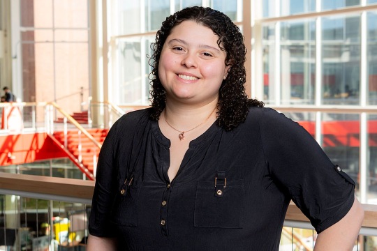 Person standing inside a modern building with large windows, wearing a navy blouse with button details.