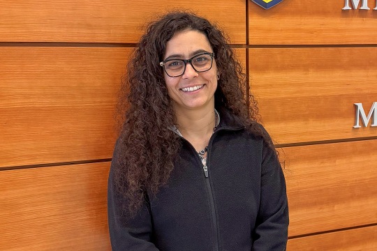 A smiling woman with long curly dark hair and black-rimmed glasses, wearing a black zip-up fleece jacket. She is standing indoors in front of a wall made of horizontal wooden panels.