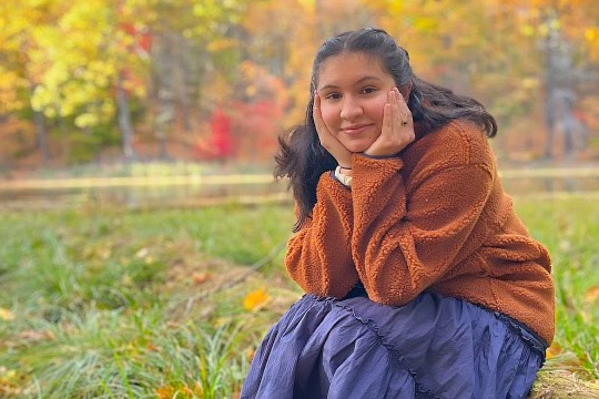 A young woman with long dark hair smiling while sitting outdoors, resting her face in both hands. She is wearing a fuzzy orange pullover and a long dark blue skirt. The background features green grass, a body of water, and trees with vibrant autumn foliage.