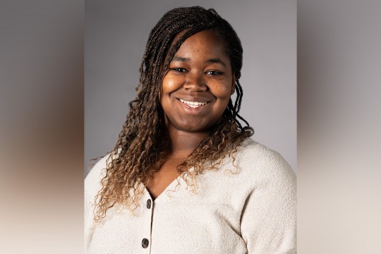 Headshot of Kierstin Delk, a smiling Black woman with long braided hair wearing a cream-colored button-up cardigan against a gray background.