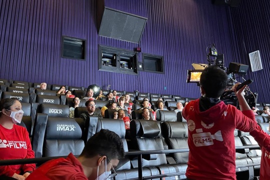 Students sitting in theater eating popcorn while being filmed. 