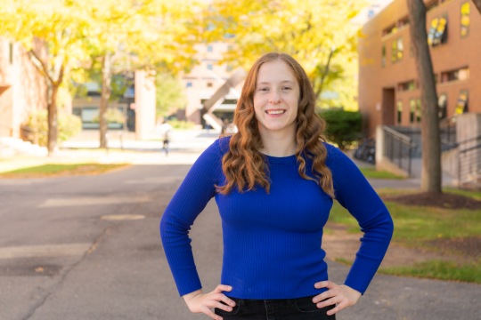 Woman with long red hair wearing a bright blue top stands with arms on hips.
