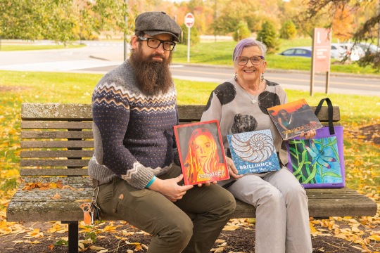 two people sitting on a bench outdoors holding a magazine and art examples.