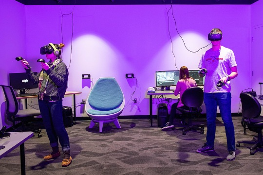 Two gaming students are wearing VR glasses and are standing in front of a purple wall. One student is sitting facing their computer screen.