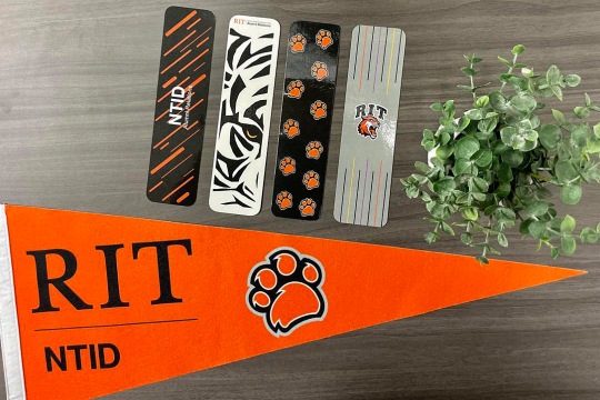 Four bookmarks laying flat. One is black with diagonal orange stripes, another is which with black graphic tiger stripes and an eye, the next is black with orange paw prints, and the last is grey with vertical lines in various colors surrounding the RIT tiger logo. Also in the photo is a pennant reading RIT NTID and a small potted plant.