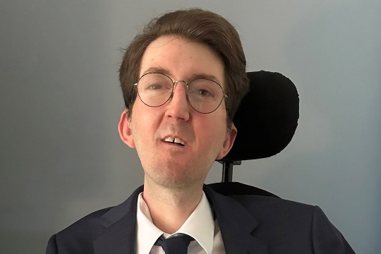 Alexander Van Hook with glasses and short brown hair, wearing a suit and tie. We see the headrest of his wheelchair behind his head.