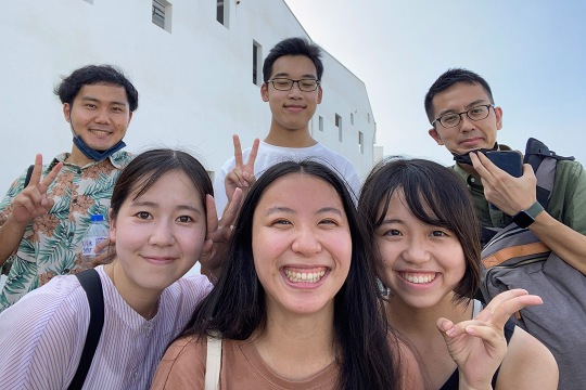 six college age students appear together for a photo in two rows of three. There is a white building in the background.