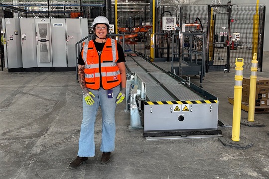 A person wearing a white hard hat, orange safety vest, black shirt, blue jeans, and yellow gloves stands in an industrial setting. The background includes machinery, control panels, and safety barriers.