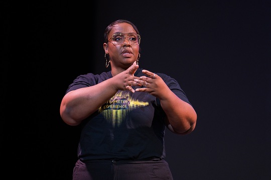 Yalanda Allgood standing on a dark stage, speaking and signing. She is wearing a black t-shirt that reads "Black Interpreter Experience."