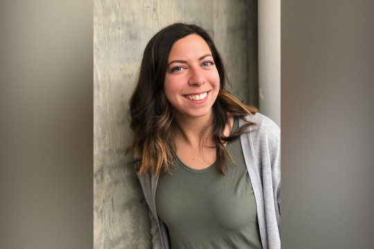 Rebecca DeSantis smiling into the camera, leaning against a textured concrete wall while wearing an olive green top and grey cardigan.