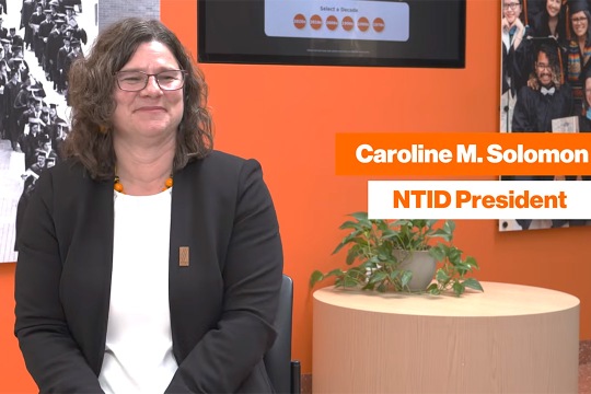 Caroline M. Solomon, NTID President, wearing glasses and a black blazer over a white top, smiling in front of a bright orange background featuring photo displays and a potted plant.