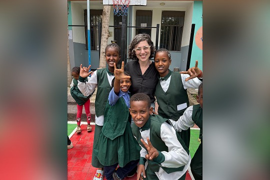 Erica Israel poses outdoors with school children in green and white uniforms, with several smiling and making the 'I love you' hand sign.