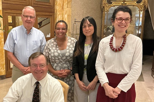 Group of five people posing indoors. They are standing and sitting in a well-decorated room with a large mirror and wooden furniture.