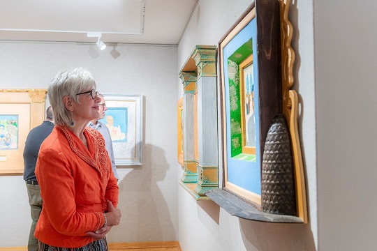Person wearing an orange jacket observing framed paintings in an art gallery, with colorful artwork displayed on light-colored walls.