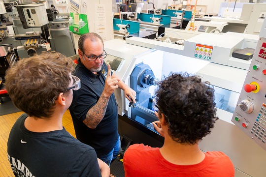 An instructor explains the operation of a CNC lathe to two students. The group is gathered closely around the machine in a well-lit industrial workshop filled with technical equipment.