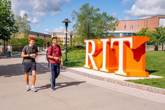 Two students with backpacks walk along a paved campus pathway on a sunny day, passing a large, orange three-dimensional "RIT" sign set on a grassy lawn with modern brick buildings in the background.