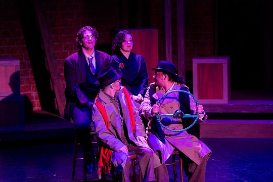 Four actors perform on a stage with purple and blue lighting during a rehearsal of "The 39 Steps." Two actors in trench coats and hats sit in the foreground, one holding a prop steering wheel, while two other actors sit behind them against a brick-patterned backdrop.
