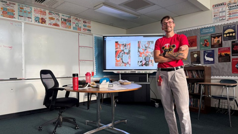 Blake Culley, an NTID graduate student, smiles while standing in a bright, modern classroom at RIT, wearing a "Be Kind" t-shirt and gesturing toward a whiteboard with educational notes.