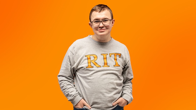 Person standing against an orange background wearing a gray sweatshirt with “RIT” printed on the front.