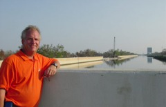 Light skinned man, light hair, orange polo shirt, arm up on cement wall, water in background.